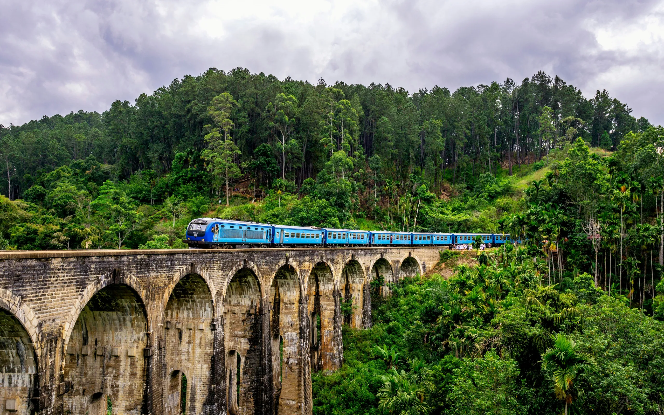 A blue train crossing the Nine Arch Bridge above the jungle canopy